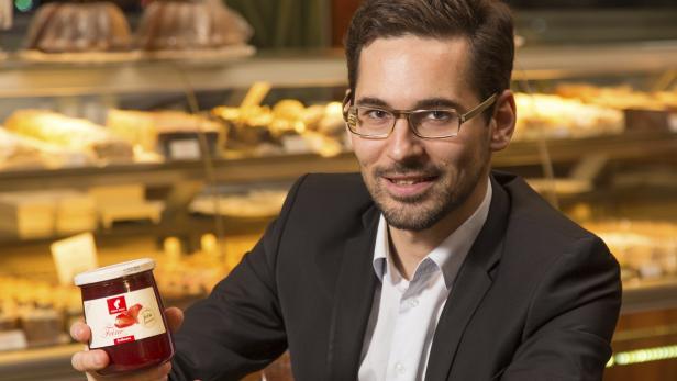 Ein Mann im Anzug präsentiert ein Glas Erdbeermarmelade vor einer Bäckereiauslage.