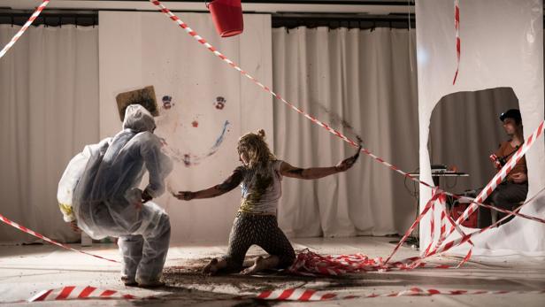 Eine Frau kniet auf dem Boden und wirft mit Erde, während eine Person im Schutzanzug daneben steht.