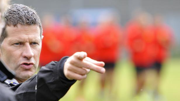 Ein Trainer gestikuliert und gibt Anweisungen, während im Hintergrund eine Gruppe von Sportlern zu sehen ist.