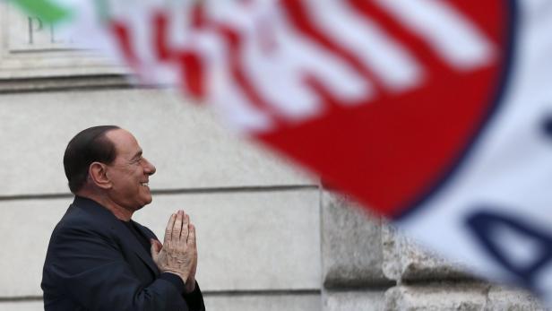Former Italian Prime Minister Silvio Berlusconi sm Silvio Berlusconi lächelt mit gefalteten Händen vor einer italienischen Flagge.