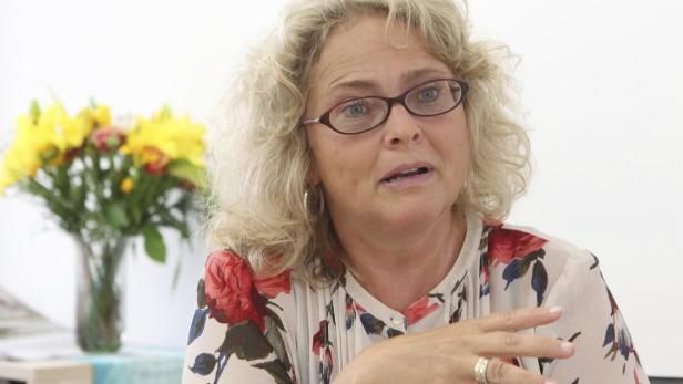 Eine Frau mit blonden Locken und Brille gestikuliert vor einem Blumenstrauß.