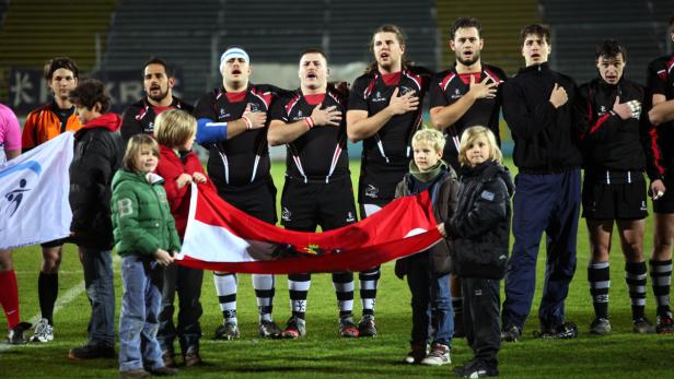 Eine Rugby-Mannschaft und Kinder halten eine rot-weiße Flagge auf einem Spielfeld.