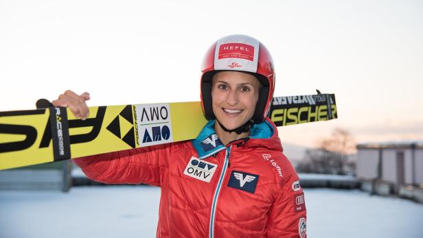 Eine Skispringerin mit Ski und Helm in roter Jacke lächelt in die Kamera.