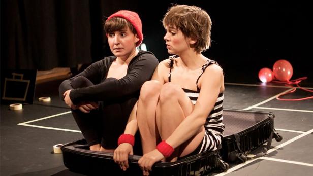 Zwei Frauen sitzen in einem Koffer auf einer Bühne.