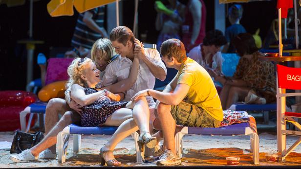 Eine lachende Frau sitzt mit zwei Männern auf einer Liege, während andere Personen im Hintergrund zu sehen sind.