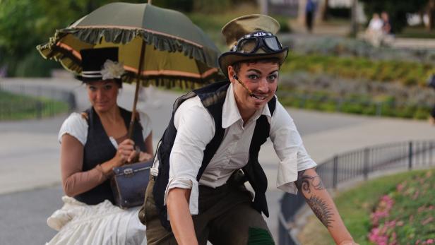 Zwei Personen im Steampunk-Stil, eine auf einem Fahrrad, die andere mit einem Sonnenschirm.