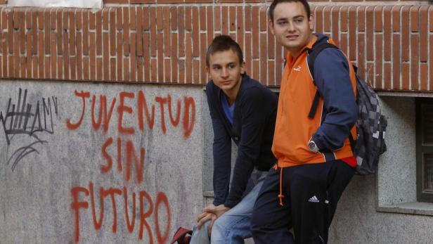 Zwei junge Männer lehnen an einer Wand mit Graffiti, auf der „Juventud sin Futuro“ steht.
