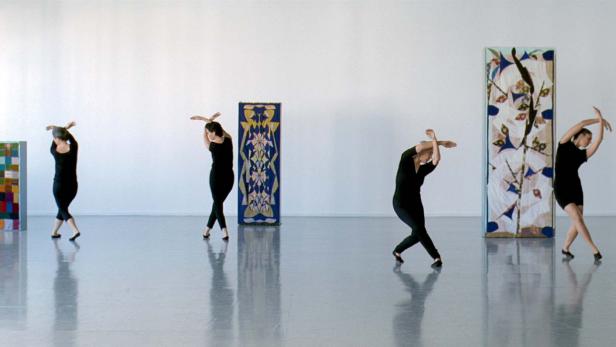 Vier Tänzerinnen in schwarzen Kleidern posieren vor abstrakten Kunstwerken in einem hellen Raum.