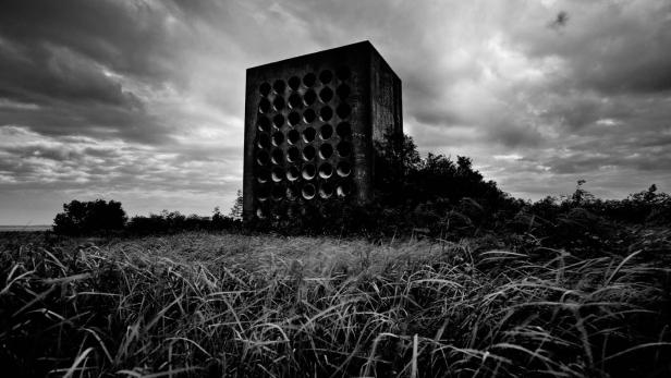 Ein würfelförmiges Gebäude mit vielen Löchern steht inmitten einer Graslandschaft.