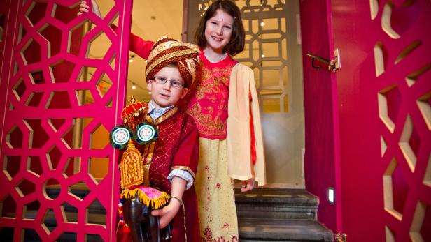 Zwei Kinder in traditioneller indischer Kleidung posieren vor einer verzierten Tür.