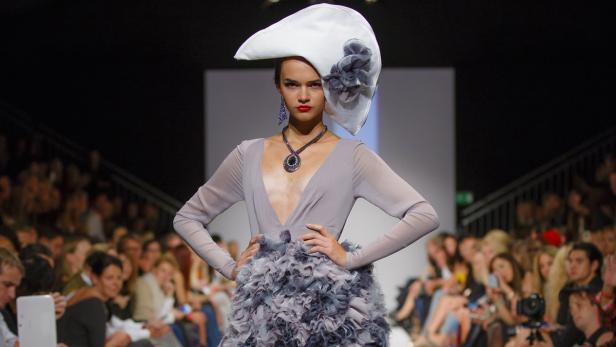 Eine Frau mit Hut und auffälligem Schmuck auf dem Laufsteg einer Modenschau.