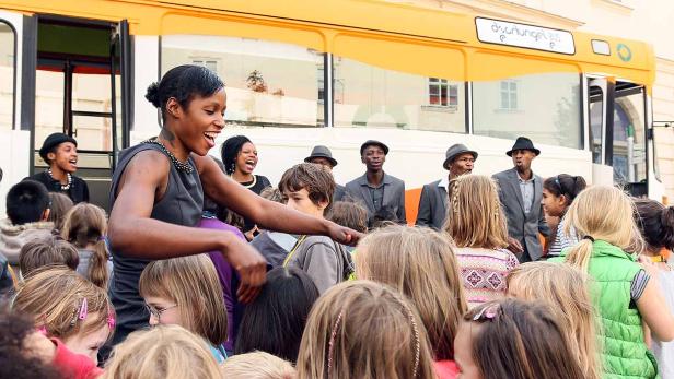 Eine Frau spricht vor einer Gruppe von Kindern vor einem gelben Bus mit der Aufschrift „Dschungel Bus“.