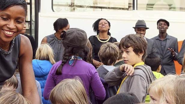 Eine lächelnde Frau interagiert mit einer Gruppe von Kindern, während eine Band im Hintergrund steht.