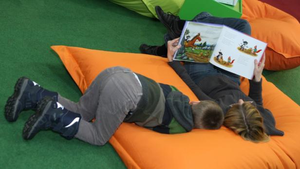 Ein Kind liegt auf einem orangefarbenen Sitzsack, während eine Frau ihm ein Buch vorliest.