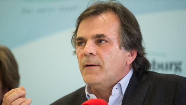 Ein Mann mit dunklen Haaren und einem dunklen Anzug spricht.