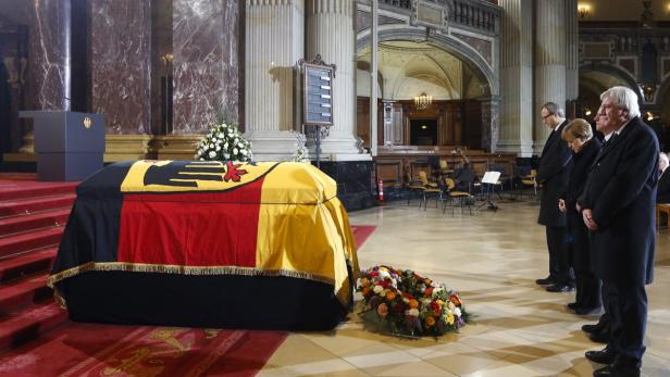 Ein mit der deutschen Flagge bedeckter Sarg in einer Kirche, umgeben von Trauergästen.