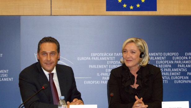 Marine Le Pen und ein Mann in einem Anzug sitzen vor einer Wand mit dem EU-Logo.