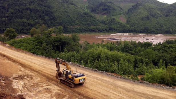 Ein gelber Bagger steht auf einer Baustelle vor einer grünen Hügellandschaft.