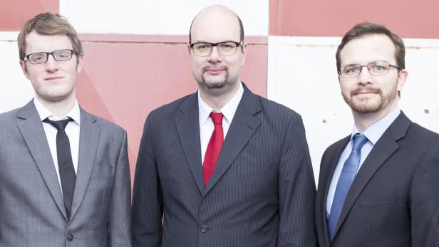 Drei Männer in Anzügen stehen vor einer rot-weißen Wand.