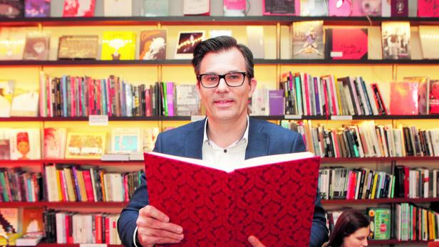 Ein Mann mit Brille liest ein rotes Buch vor einem Bücherregal.