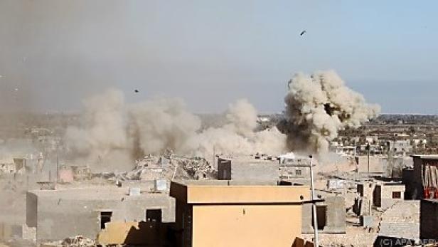 Eine Explosion inmitten einer zerstörten Stadtlandschaft hüllt Gebäude in Staub.