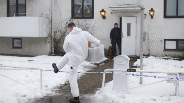 Ein Mann in Schutzanzug trägt eine Kiste über ein Absperrband vor einem Haus.