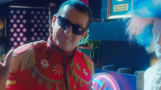 Ein Mann mit Sonnenbrille und roter Uniform steht neben einer Büste mit blauer Zuckerwattefrisur.