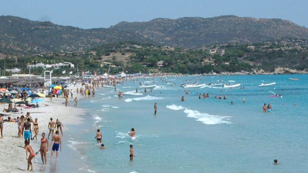Ein belebter Strand mit vielen Menschen, Sonnenschirmen und Booten im türkisfarbenen Wasser.