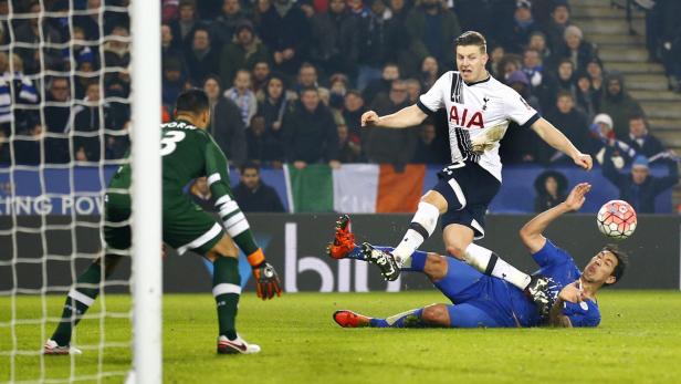 Ein Fußballspieler springt über einen am Boden liegenden Gegenspieler, während der Torwart zusieht.