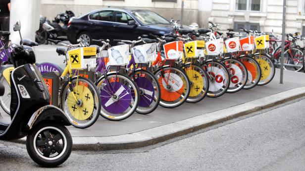 Ein schwarzer Motorroller und eine Reihe von bunten Leihfahrrädern stehen am Straßenrand.