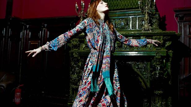 Eine Frau in einem bunten Kleid steht vor einem verzierten, grünen Kamin.