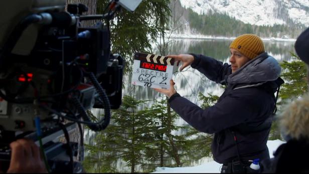 Ein Mann mit Mütze hält eine Filmklappe für den Film „Spectre“ vor einer winterlichen Landschaft.