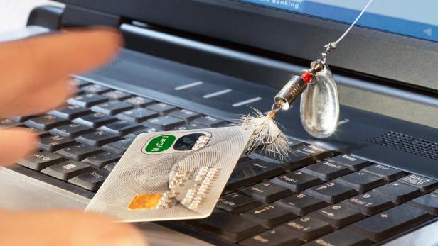 Eine Kreditkarte und ein Angelhaken liegen auf der Tastatur eines Laptops, der Online-Banking anzeigt.