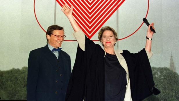 Eine Frau mit Mikrofon und ein Mann mit Fliege posieren vor einem Logo.