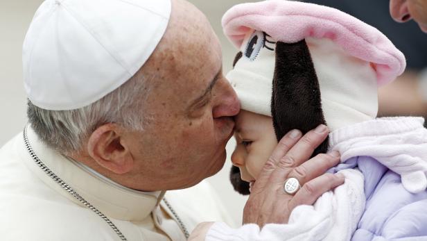 Papst Franziskus küsst ein Baby mit einer Mütze in Hundeform auf die Stirn.