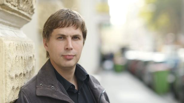 Ein Mann mit braunen Haaren lehnt an einer Wand.