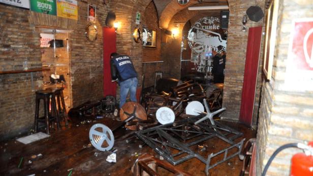Ein Tatort in einer Bar mit umgestürzten Stühlen und Tischen, untersucht von der Polizei.