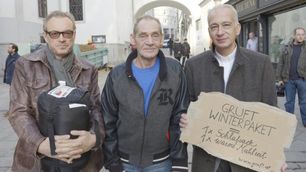Drei Männer posieren mit einem Schlafsack und einem Schild für das „Gruft Winterpaket“.