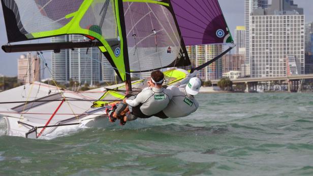 Zwei Segler auf einem kleinen Segelboot vor der Skyline einer Stadt.