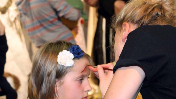 Ein Mädchen mit Blumen im Haar wird geschminkt, um eine Wunde auf der Stirn darzustellen.