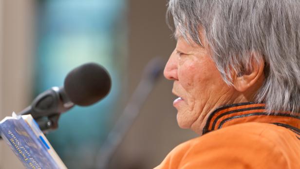 Eine ältere Frau liest aus dem Buch „Galsan Tschinag: Gold und Staub“ vor einem Mikrofon.