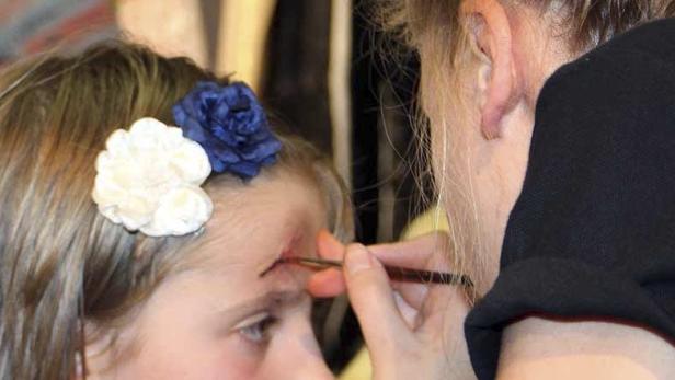 Einem Mädchen mit Blumen im Haar wird ein künstliche Wunde ins Gesicht geschminkt.