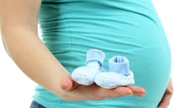 Eine schwangere Frau hält blaue Babyschuhe in ihrer Hand.