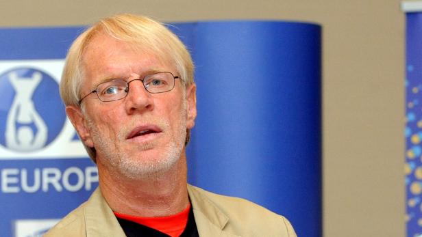 Ein Mann mit blonden Haaren und Brille vor einem blauen Hintergrund.
