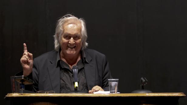 Ein Mann mit weißem Haar gestikuliert während einer Rede hinter einem Rednerpult.