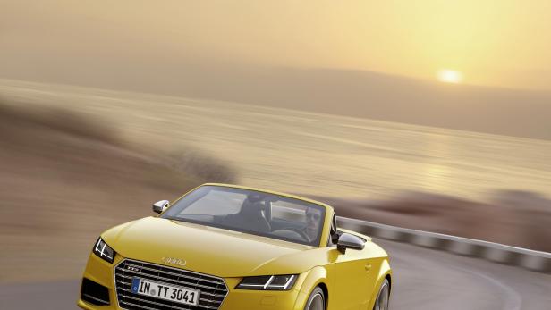 Ein gelbes Audi TT Cabriolet fährt auf einer Küstenstraße in Richtung Sonnenuntergang.