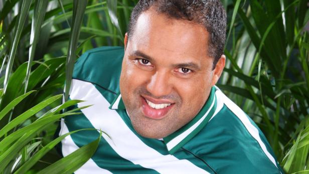 Ein lächelnder Mann mit einem grün-weiß gestreiften Hemd steht zwischen tropischen Pflanzen.