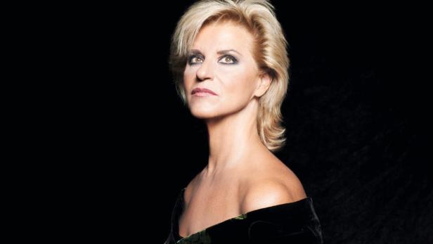 Eine Frau mit blonden, kurzen Haaren und dunklem Kleid vor schwarzem Hintergrund.