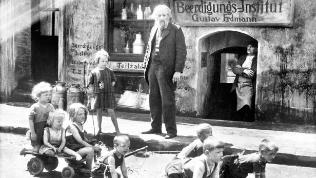 Kinder spielen vor dem „Beerdigungs-Institut Gustav Erdmann“ in einer Straßenszene.