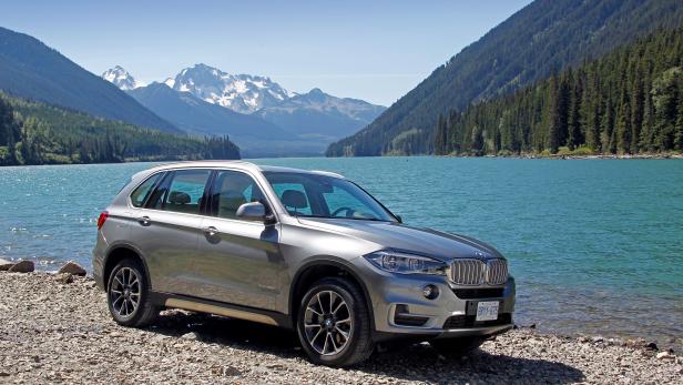 Ein silberner BMW X3 steht an einem See mit Bergen im Hintergrund.